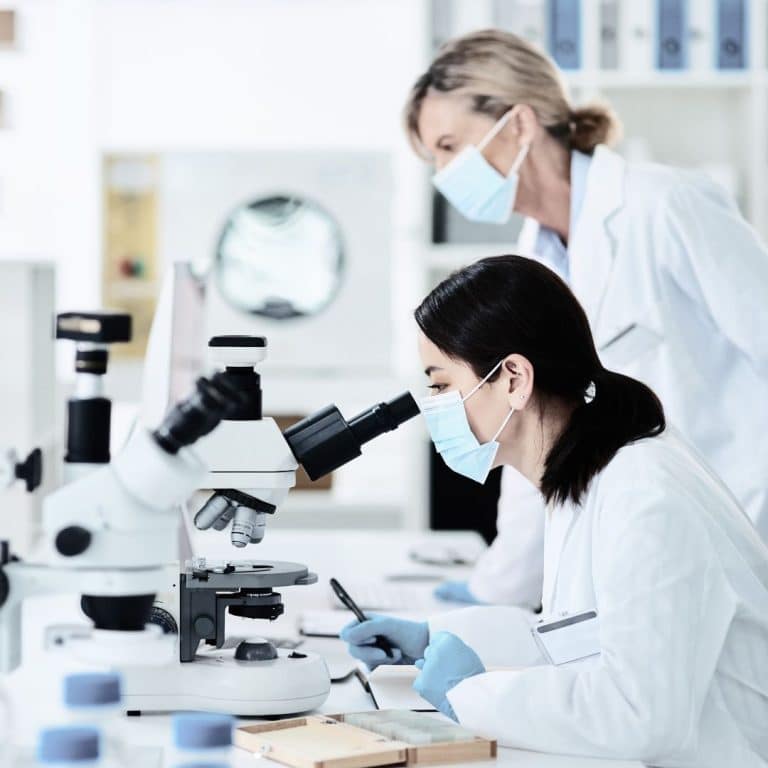 women in white lab coats and masks looking through a microscope